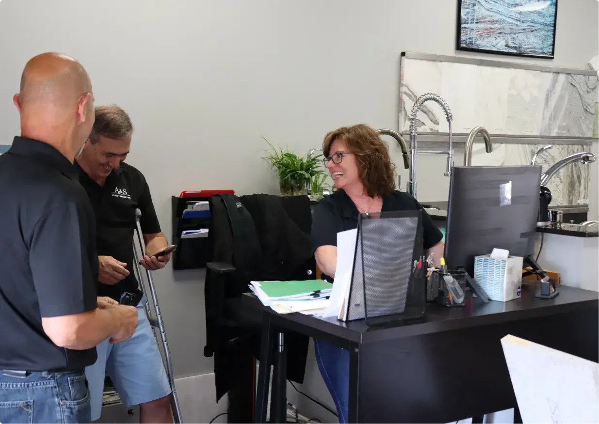A friendly interaction between three team members inside the A&S Stone Fabricators office. A woman sitting at a desk is smiling while talking to two standing men, one of whom is using crutches.