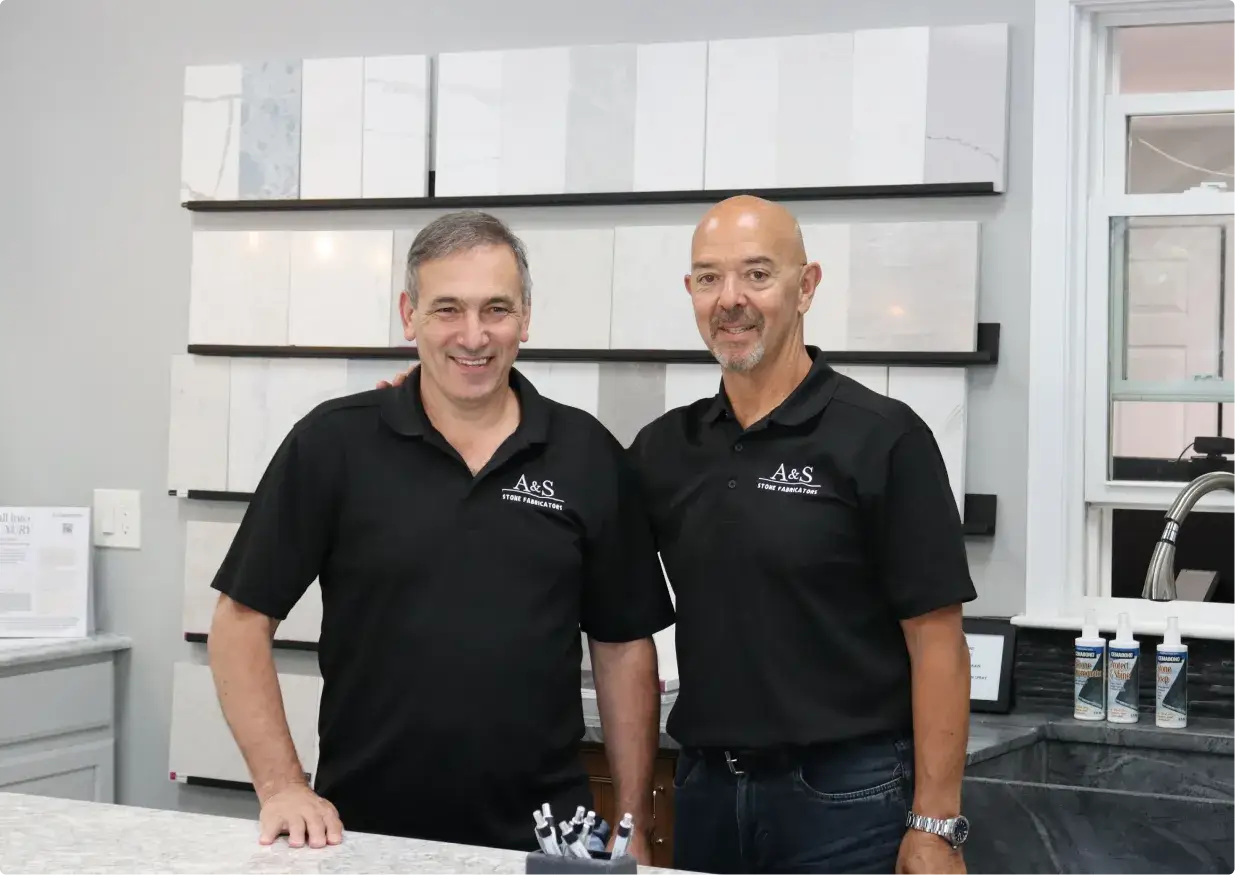 Two male team members of A&S Stone Fabricators standing together, smiling in a showroom with various stone samples displayed on shelves behind them.